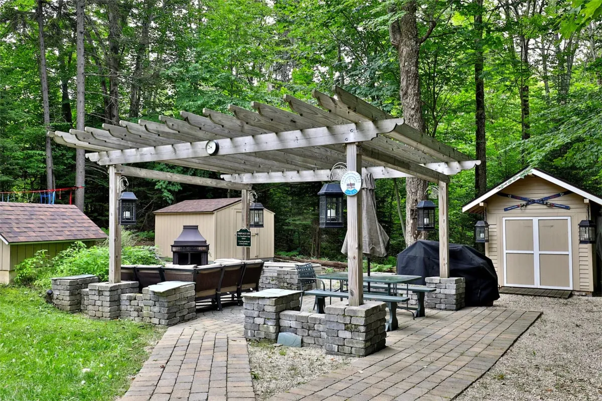 An inviting outdoor space featuring a pergola with comfortable seating, a grill, and outbuildings, set up for group gatherings or relaxed outdoor meals. The area is designed for communal activities, with ample room for multiple guests to socialize, dine, or enjoy the natural setting.