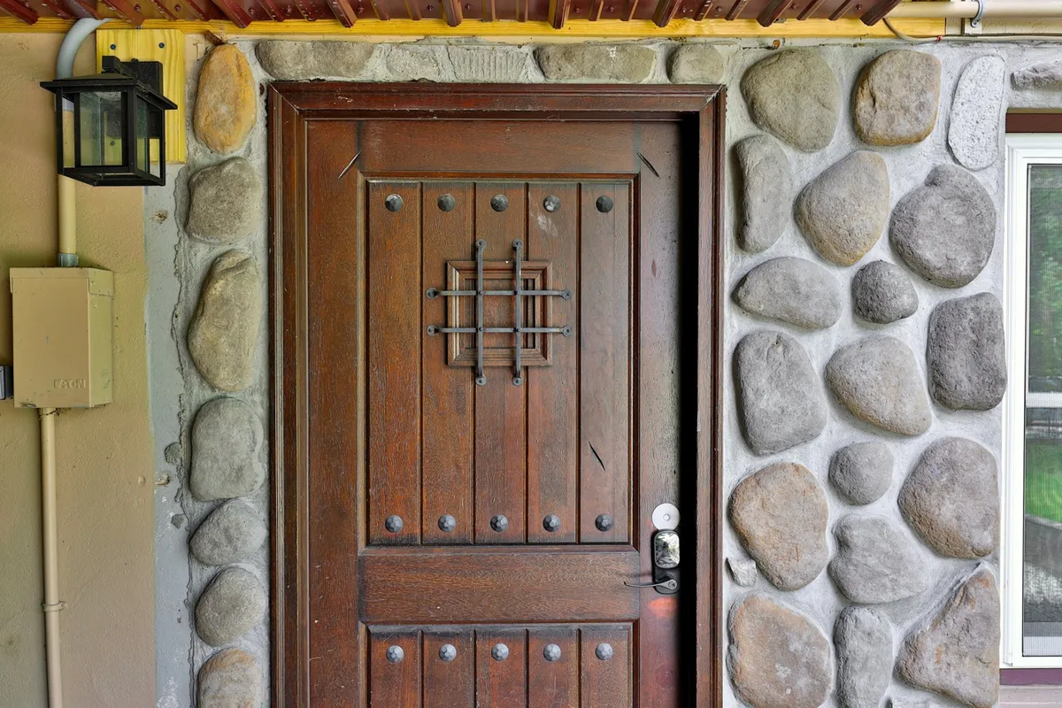 A close-up of a warmly lit wooden door set into a textured stone wall, illuminated by a lantern. The view suggests the transition from the lively outdoors into a cozy, private space.