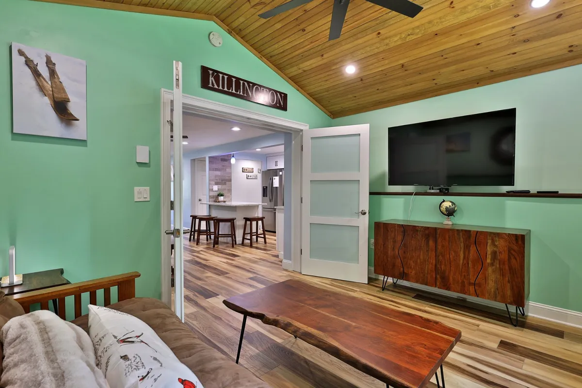 A den-like area with a wooden coffee table, a wall-mounted TV, artistic wall decor, and an open doorway leading toward the kitchen. The room feels versatile and intimate, ideal for reading, playing games, or transforming into an extra sleeping area.