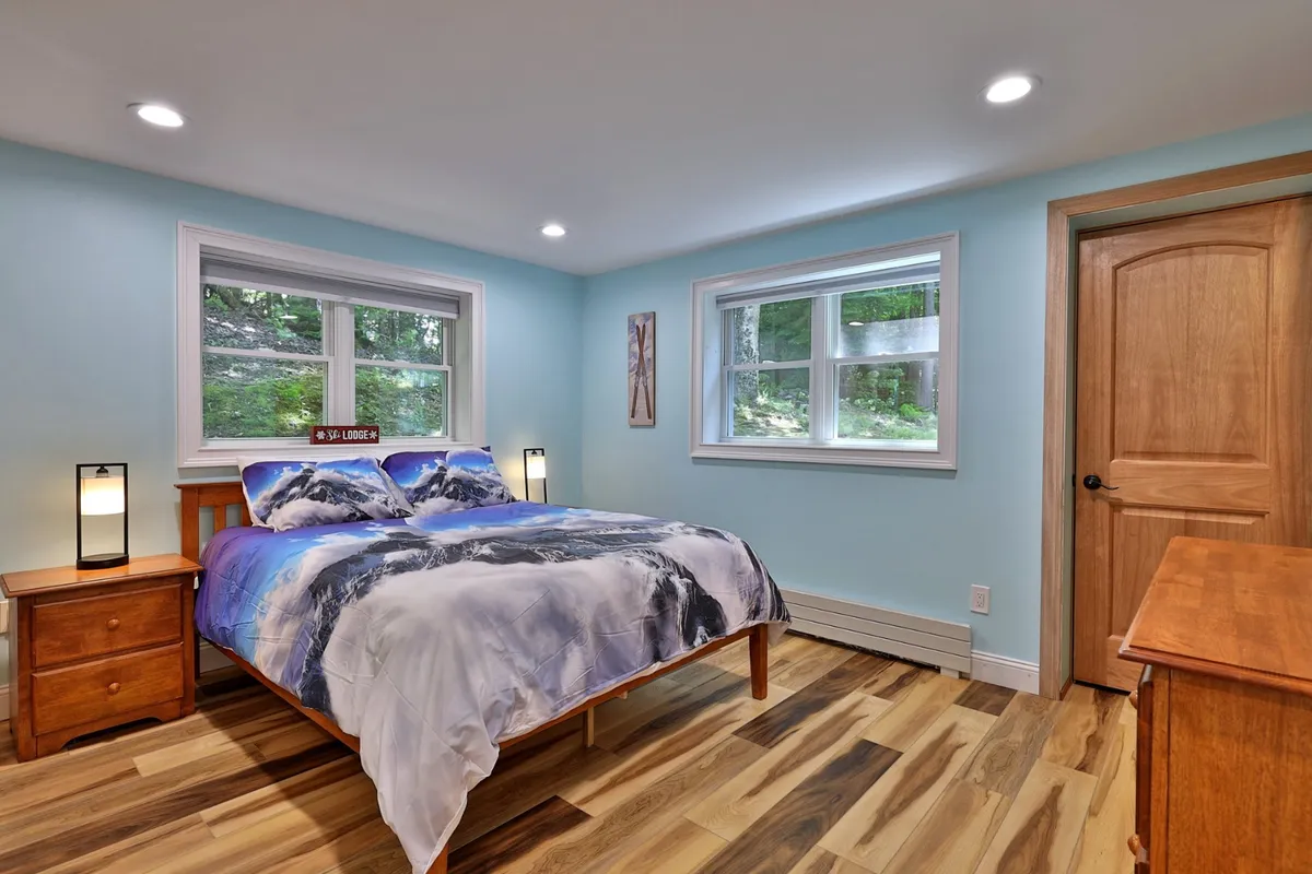 A serene bedroom featuring mountain-themed bedding, a wooden nightstand, and large windows that allow natural light to flood the space. A wooden door is visible, adding to the room's rustic charm.