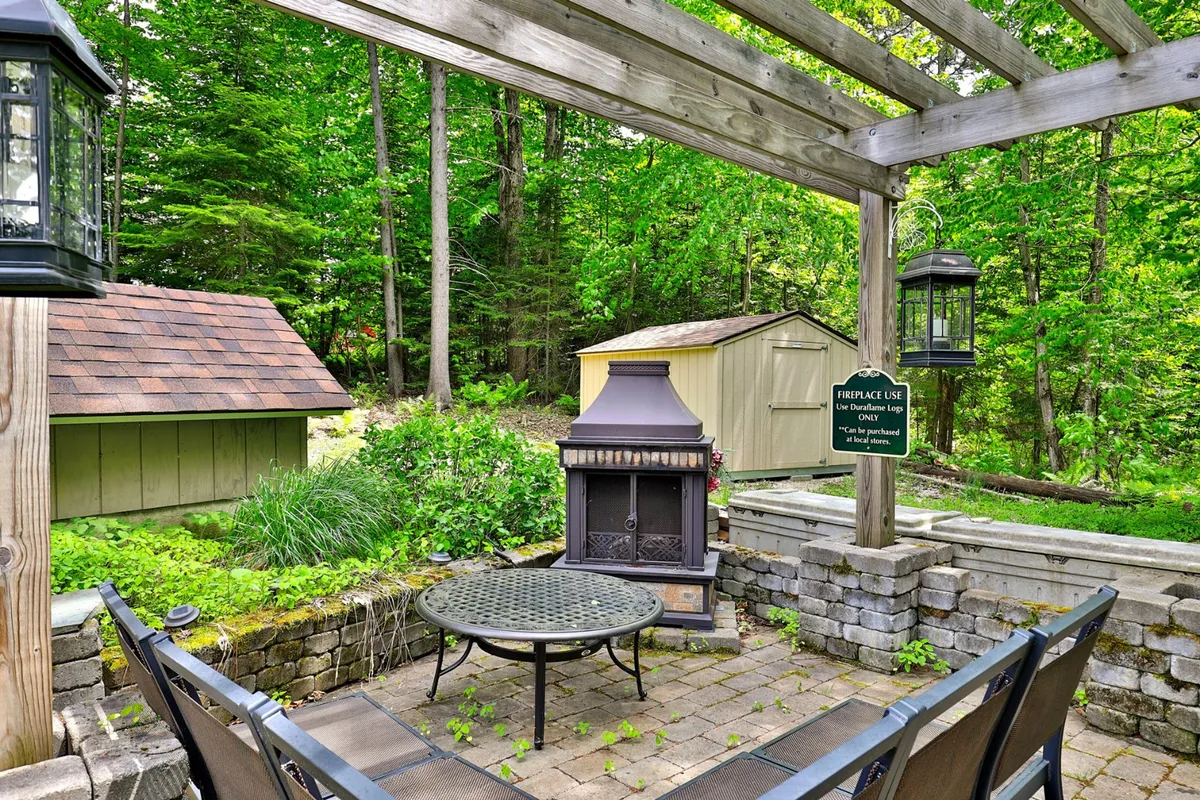 A cozy outdoor patio with a fireplace, dining table, pergola, and surrounding greenery. The space is intimate yet open, suggesting fireside chats, outdoor meals, and a tranquil setting for socializing. The greenery adds to the all-seasons appeal, making it suitable for both warm and cool weather activities.