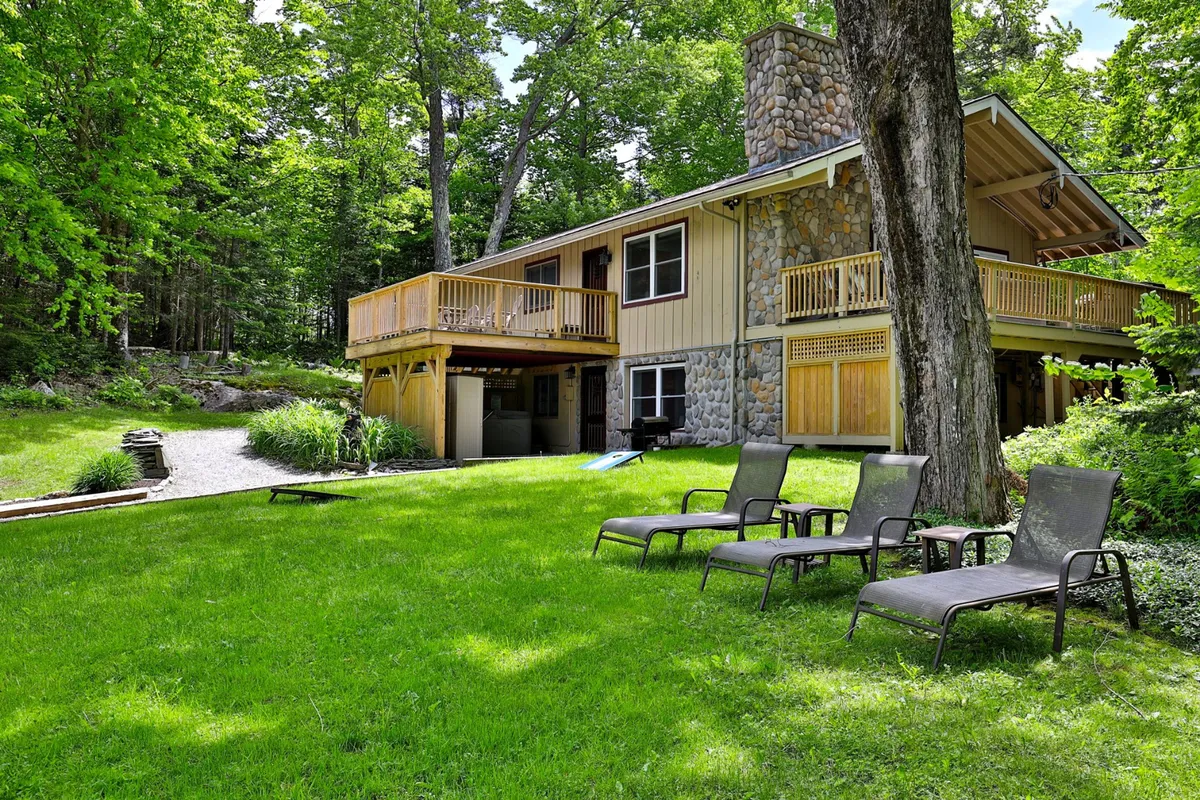 A wide view of the home's exterior shows the house with its stone chimney, a welcoming outdoor seating area, and a lush green lawn. The scene is bright and open, suggesting space for play and relaxation. The balcony and lawn both suggest opportunities for family-friendly activities in an inviting, natural setting.