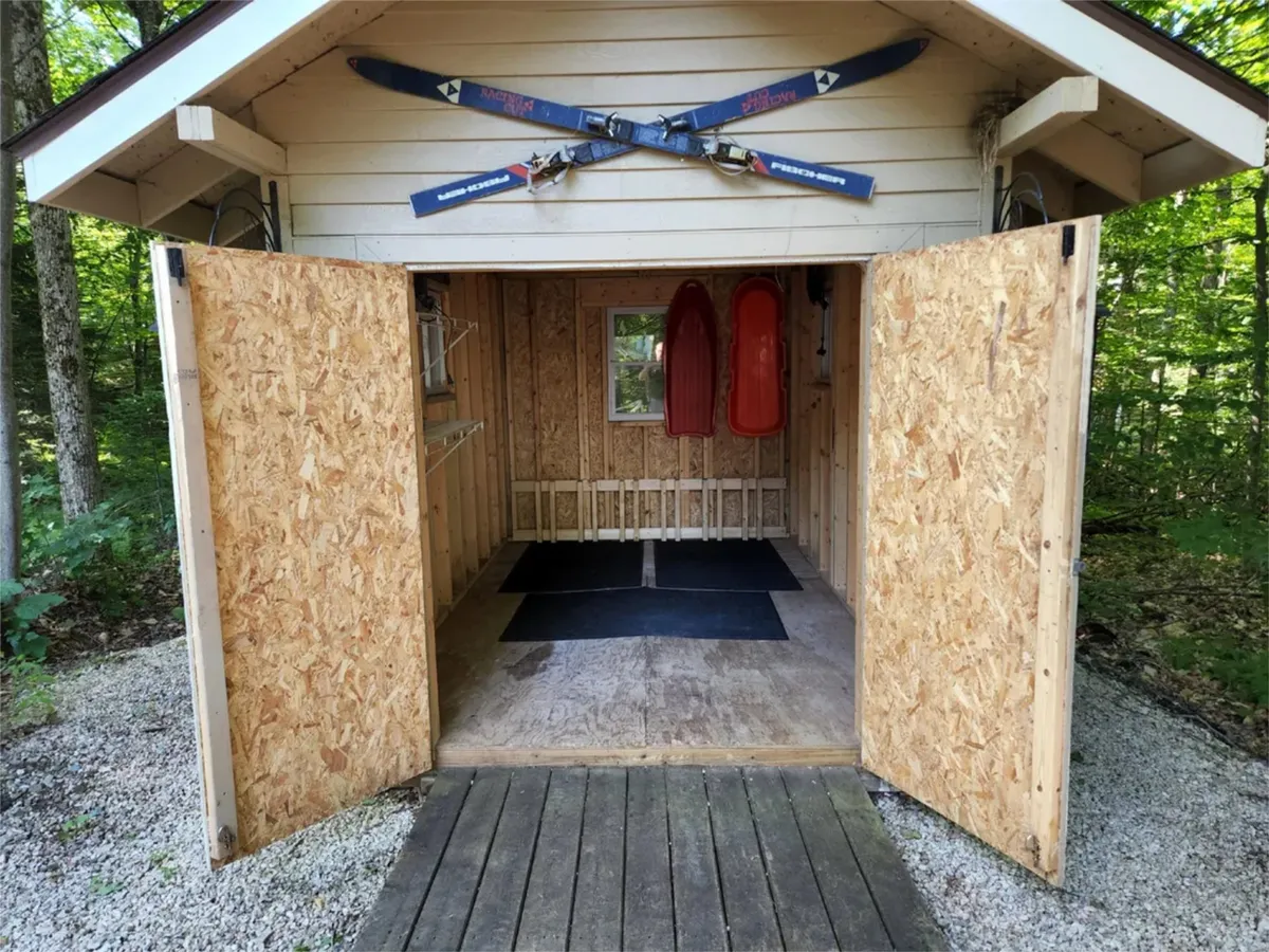 A cozy mudroom or entryway featuring crossed skis mounted on the wall, two red sleds leaning nearby, and open double doors inviting in the winter air. The scene evokes memories of prepping for a day on the slopes or storing gear after a snowy adventure.