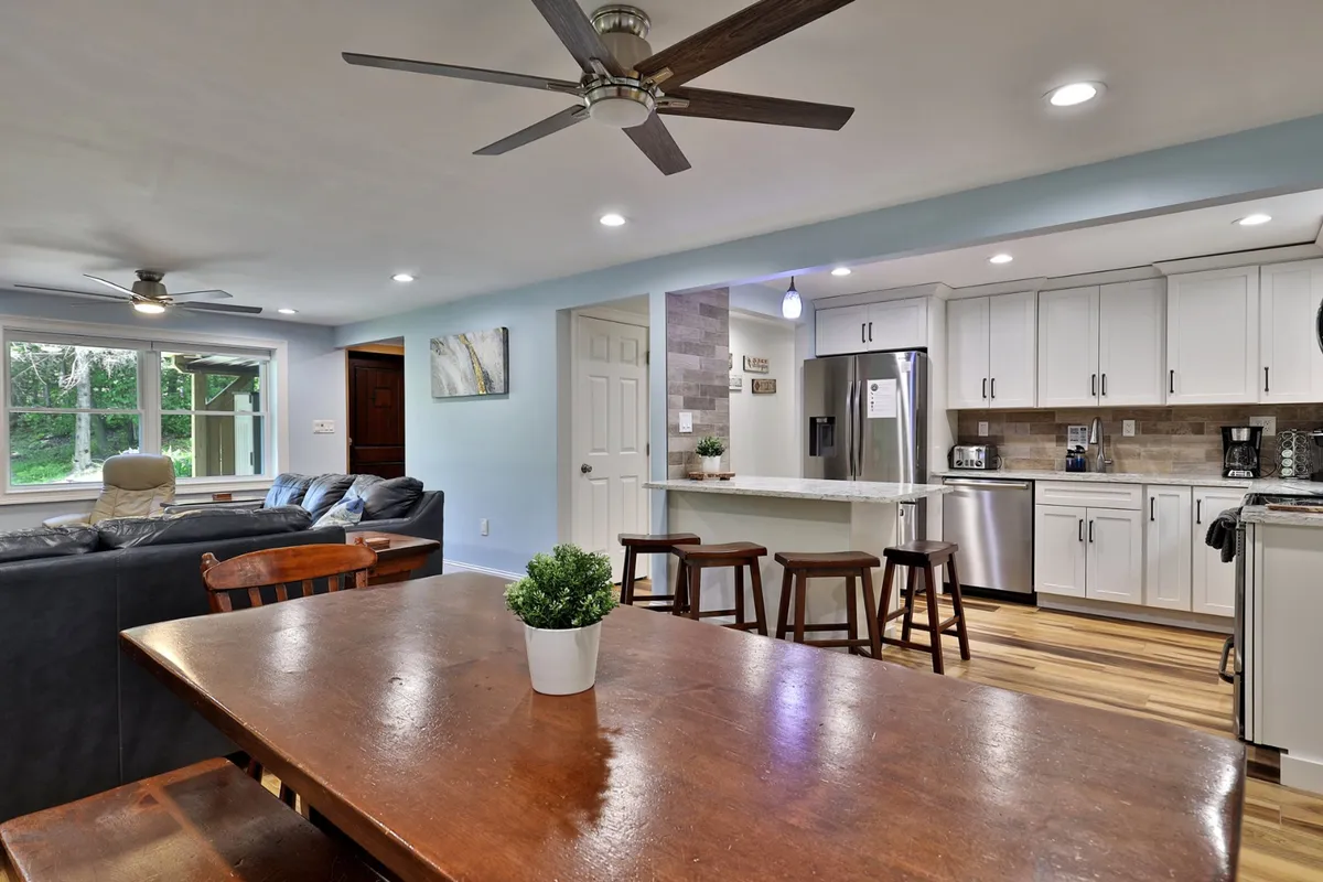 This image provides a wide view of the kitchen and dining area, featuring a spacious dining table, a kitchen island, a sofa, and large windows that let in plenty of natural light. The granite countertops and stainless steel appliances are visible, highlighting the kitchen’s modern amenities. The space seamlessly connects the areas for cooking, dining, and relaxing.