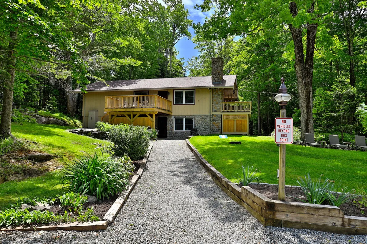A welcoming view of the house featuring a front path, a balcony ideal for sunset views, and a prominent sign, all framed by lush greenery. The scene radiates a sense of arrival and is inviting for groups or couples returning from an evening out.