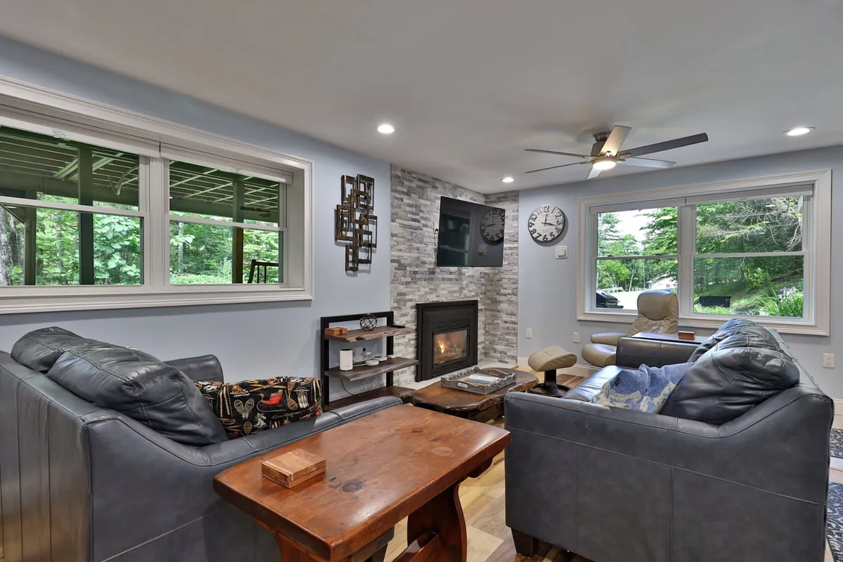The image features a cozy, freshly renovated living space with a crackling fireplace as the focal point, surrounded by comfortable seating and tasteful wall decor, all set against the backdrop of large windows that hint at the tranquil wooded surroundings.