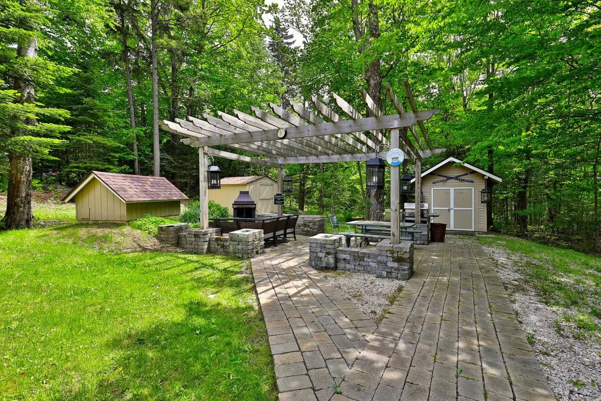 A comfortable pergola area with a stone seating arrangement, grill, and small shed set against a green backdrop. The shed hints at secure storage for bikes and gear, while the grill and seating suggest shared barbecues and group activities. The atmosphere is relaxed and communal, fitting for family fun.