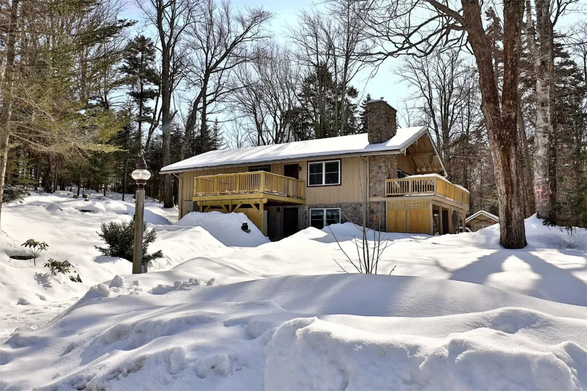 A picturesque winter scene with the house nestled among snow-covered trees, the ground blanketed in fresh white snow. The porch and deck are visible, inviting guests to enjoy the crisp mountain air after a day on the slopes.