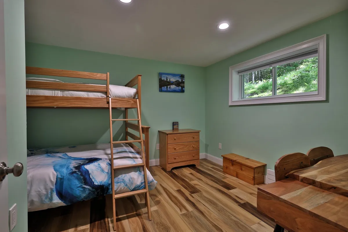 A cozy bunk room with a wooden bunk bed, a dresser, a window with a view, and an art piece on the wall. The space feels inviting and playful, ideal for families and children to relax or play.
