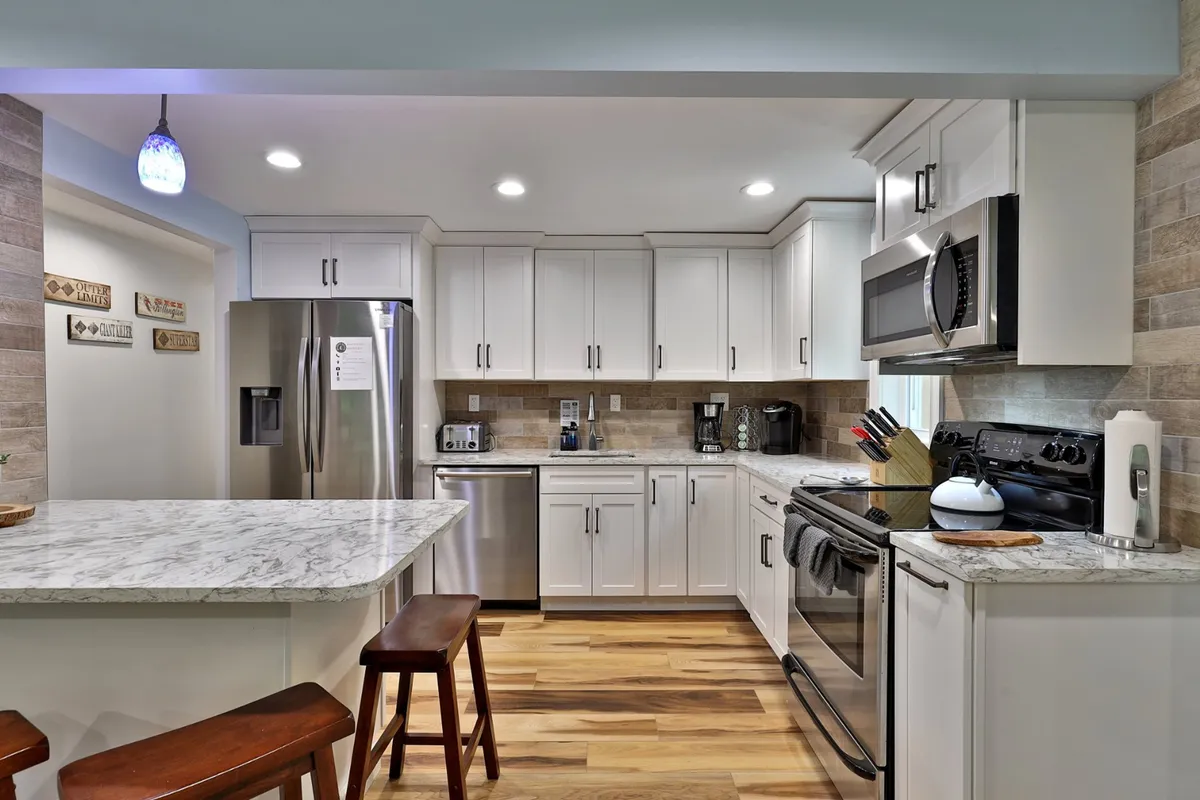 A modern kitchen with a marble countertop, stainless steel appliances, and wooden bar stools, designed for social cooking and communal dining. The space is open and welcoming, perfect for preparing shared meals or hosting group breakfasts.