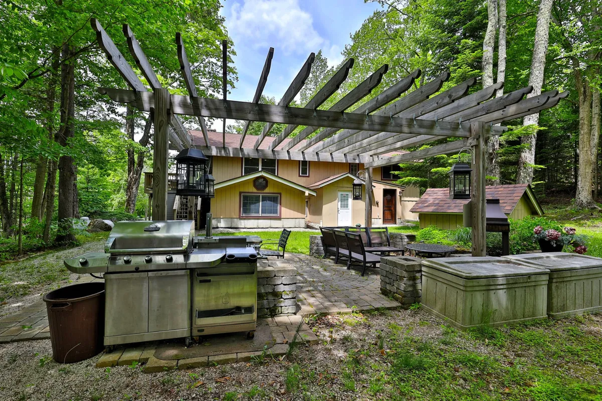 An inviting outdoor kitchen area featuring a grill under a pergola with a cozy seating arrangement, all set amid green surroundings. The space is designed for outdoor dining, relaxation, and social gatherings, with plenty of natural light and comfort.